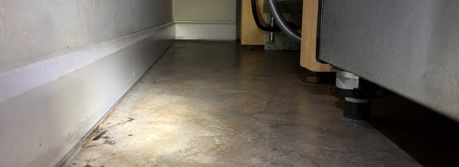 Luce di una torcia che illumina lo spazio stretto dietro un mobile da cucina, con tracce scure sul pavimento